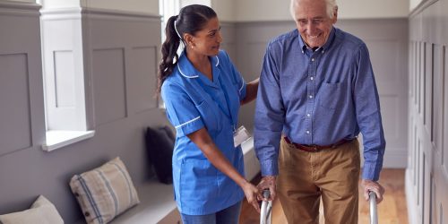 senior man at home using walking frame being helped by female ca