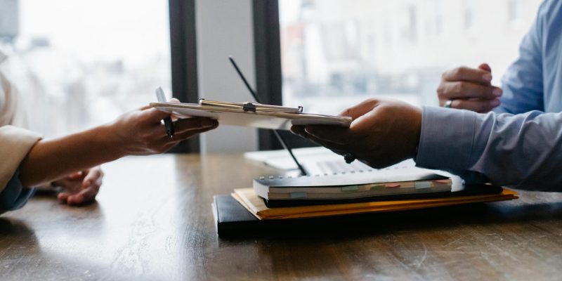 Crop anonymous ethnic woman passing clipboard to office worker with laptop during job interview